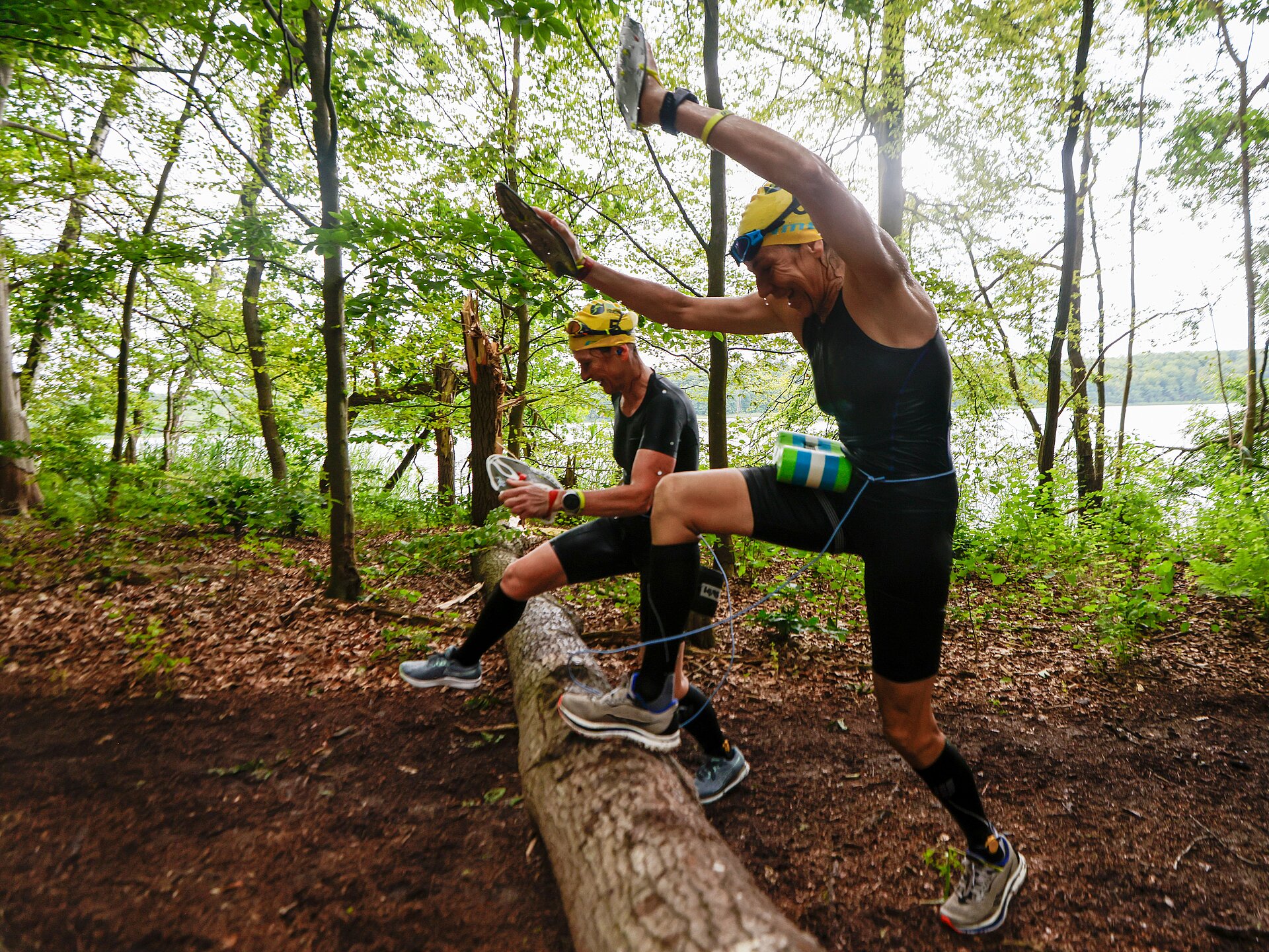 SwimRun: Female runners jump over a tree trunk in the forest © SCC EVENTS / Jean-Marc Wiesner